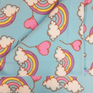 Può includere: Una coperta in pile azzurro con un motivo ripetuto di arcobaleni, nuvole bianche e palloncini rosa a forma di cuore. Gli arcobaleni hanno strisce gialle, arancioni e viola. La coperta è piegata.