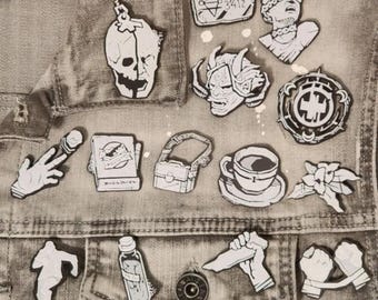 May include: A collection of white and black enamel pins with various designs, including a skull, a bust, a cup of coffee, and a hand holding a knife. The pins are arranged on a gray denim jacket.