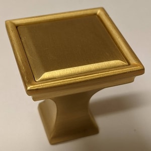 May include: A gold square cabinet knob with a raised, beveled top and a decorative base.