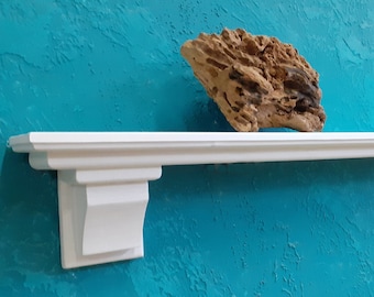 Puede incluir: Estante blanco con molduras decorativas en una pared turquesa. El estante está montado en la pared con soportes. Un trozo de madera a la deriva está sentado en el estante.