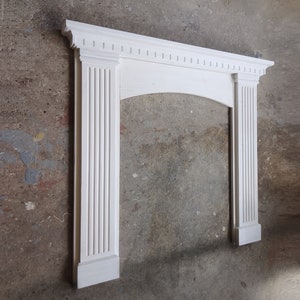 Könnte beinhalten: Ein weißer Kaminsims mit klassischem Design. Der Kaminsims hat kannelierte Säulen auf beiden Seiten der Öffnung und eine dekorative Oberseite mit kleinen rechteckigen Details. Die bogenförmige Öffnung wird von der Struktur des Kaminsims eingerahmt.