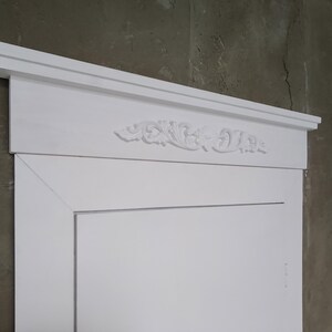 Puede incluir: Un marco de puerta blanco y ornamentado con una moldura superior decorativa. El marco presenta un diseño floral tallado y detallado en el centro de la sección superior. El marco de la puerta está pintado de blanco y colocado contra una pared gris texturizada.