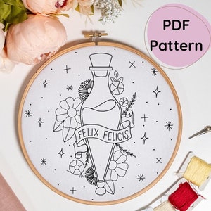 May include: A black and white embroidery pattern of a potion bottle with the text "Felix Felicis" and floral accents. The pattern is on a white fabric hoop.