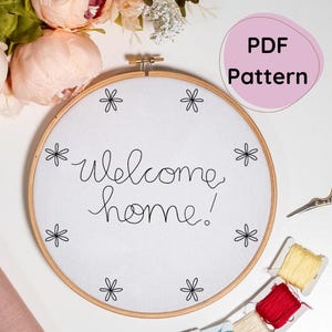 May include: A white embroidery hoop with the text "welcome home!" stitched in black thread. The hoop is surrounded by small black flower designs.