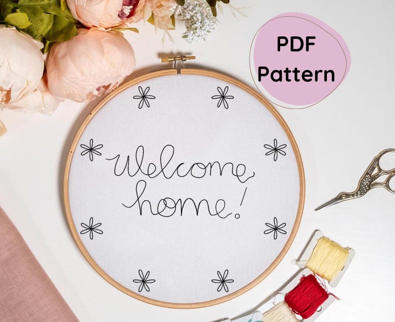 May include: A white embroidery hoop with the text "welcome home!" stitched in black thread. The hoop is surrounded by small black flower designs.