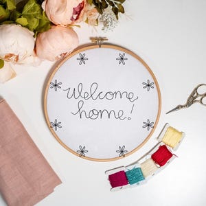 May include: A white embroidery hoop with the words "welcome home!" stitched in black thread. The hoop is surrounded by pink flowers, embroidery floss, and a pair of silver scissors.