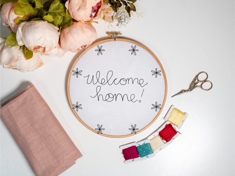 May include: A white embroidery hoop with the words "welcome home!" stitched in black thread. The hoop is surrounded by pink flowers, embroidery floss, and a pair of silver scissors.