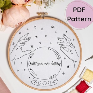 May include: A black and white line drawing embroidery pattern of a hand reaching towards a crystal ball with the text "Craft your own destiny" inside the ball. The pattern is on a white background and surrounded by a wooden embroidery hoop.