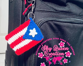 May include: A crocheted Puerto Rican flag keychain in red, white, and blue hangs from a black suitcase. The keychain features the flag's design with a white star on a blue triangle. The suitcase has multiple zippers.