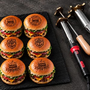 May include: Three grilled meat patties and six assembled cheeseburgers on a black slate board. The patties and buns are branded with text including Michelle Burger Co, Anchor, and Lunch Deli. Three metal branding irons with wooden handles sit nearby.