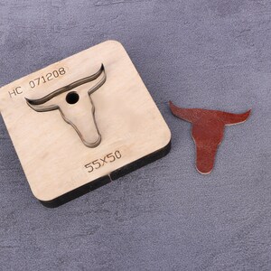 May include: A wooden leather craft punch with a bull's head design. The punch is labeled "HC 071208" and "55X50". A brown leather cutout of a bull's head is shown next to the punch.