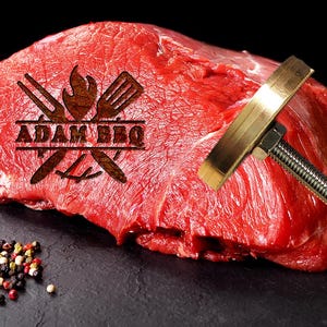 Puede incluir: Un gran corte de carne roja cruda con el texto "ADAM BBQ" marcado en su superficie. Un termómetro de carne metálico está insertado en la carne. Un pequeño montón de granos de pimienta y un tomate están en primer plano.