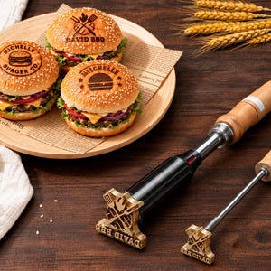 May include: Two metal branding irons with wooden handles, one large and one small, featuring a crossed spatula and fork design. Next to them are three sesame seed buns on burgers, each branded with text: Michelle Burger Co, David BBQ, and Michelle.