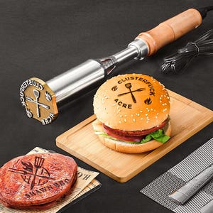 Könnte beinhalten: Ein Brenneisen mit Holzgriff und Metallstempel, neben einem Hamburger und einem Steak, beide mit Logo und Text versehen. Der Hamburger liegt auf einem Holzschneidebrett. Das Steak liegt auf einem Stück Zeitung.