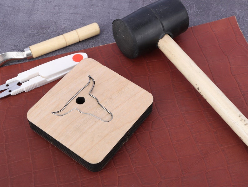 May include: A wooden leather craft punch shaped like a bull's head with a metal cutting die. A rubber mallet and a pair of scissors are also in the image.