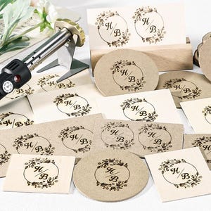 May include: A wood-burning tool with a gold tip and black handle, alongside various paper and wood pieces. The pieces are adorned with a floral wreath design and the letters "H/B". The items are in shades of beige and brown.