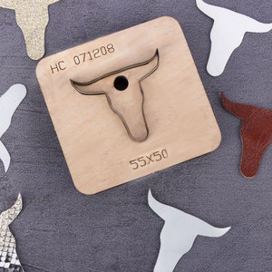 May include: A wooden craft die with a bull's head design. The die is labeled "HC 071208" and "55X50". There are several cut-out bull's heads made from different materials, including leather, glitter, and fabric.