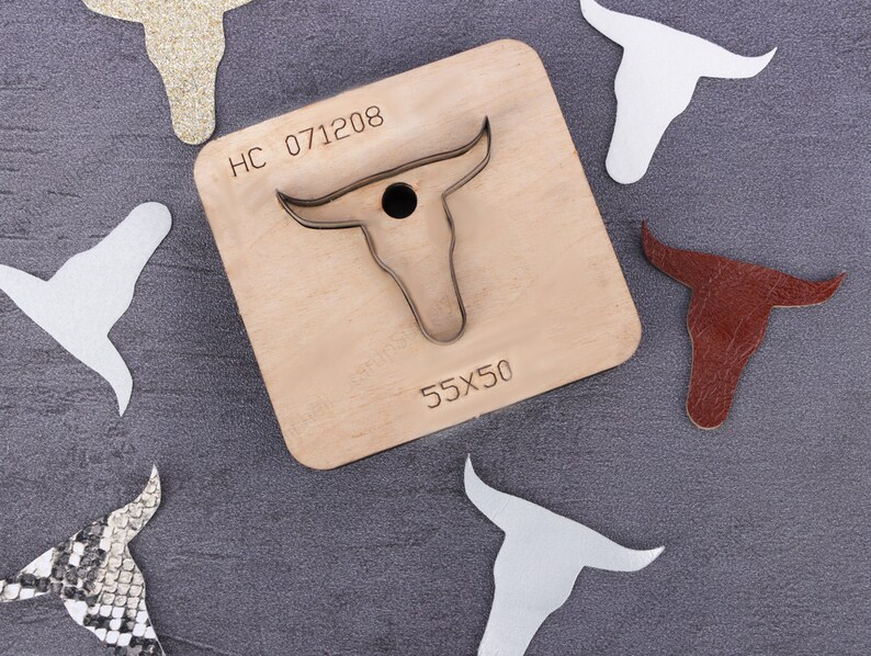 May include: A wooden craft die with a bull's head design. The die is labeled "HC 071208" and "55X50". There are several cut-out bull's heads made from different materials, including leather, glitter, and fabric.