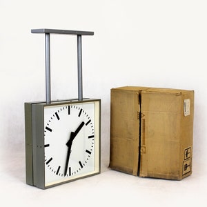Könnte beinhalten: Eine graue quadratische Uhr mit schwarzen Zeigern und einem weißen Zifferblatt. Die Uhr hängt an einem Metallrahmen und steht neben einem braunen Karton.