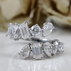 May include: A silver ring featuring a collection of clear gemstones in diverse shapes, such as pear, emerald, and round cuts. The ring's design is open, with the gemstones arranged in a flowing, asymmetrical pattern.