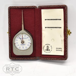 May include: A silver hand grip strength tester with a dial that reads up to 150 grams. The tester is in a brown leather case with the words "DYNAMOMÉTRE" and "DYNAMOMETER" printed on the inside. The case also has the words "GROUPE CARPANO" and "CLUSES (Hte-Savoie)" printed on the inside.