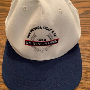 Op de afbeelding: Een witte baseballcap met een marineblauwe klep. De cap heeft geborduurde tekst met de tekst "Des Moines Golf & CC 1999 U.S. Senior Open" in rood, zwart en grijs. De cap is gemaakt van een zachte, duurzame stof.