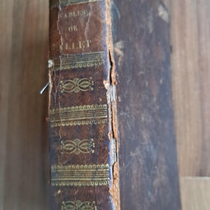 May include: Antique book with a worn, brown leather cover. The spine features gold lettering and decorative gold accents. The title "FABLES OF GILLET" is visible. The book shows signs of age and wear.