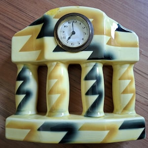 May include: A vintage ceramic clock in a cream, yellow, and black geometric design. The clock face is round with a brass-colored frame and black numerals. The clock has a decorative architectural shape with columns.