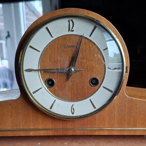 May include: A vintage wooden mantel clock with a light brown finish. The clock face is white with black numerals and hands, and a gold-coloured trim. The clock has a curved top and a rectangular base.