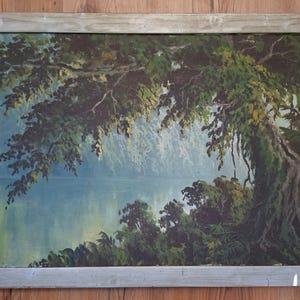 May include: A rectangular landscape painting in a weathered light gray wooden frame. The artwork depicts a serene lake scene with a large, dark green tree extending over the water, surrounded by lush foliage and a soft, misty blue background.