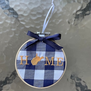 May include: A blue and white checkered fabric hoop ornament with the word "HOME" embroidered in gold thread. The state of West Virginia is also embroidered in gold thread in the center of the word "HOME".