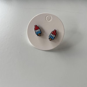 Pode incluir: Brincos em forma de picolé. Cada brinco apresenta uma parte superior vermelha, um centro branco e uma parte inferior azul, com um palito de madeira. Os brincos são exibidos em um cartão circular branco.