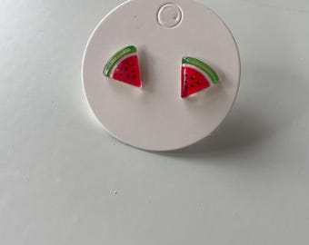 Könnte beinhalten: Ein Paar Ohrringe in Form von Wassermelonenscheiben. Jeder Ohrring hat ein rotes Dreieck mit schwarzen Samendetails und einer grünen Schale. Die Ohrringe werden auf einer weißen Ohrringkarte präsentiert.
