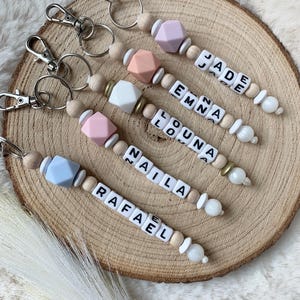 Peut inclure: Cinq porte-clés personnalisés avec des perles en bois et des lettres en silicone épellant les noms JADE, EMMA, LOUNA, NAILA et RAFAEL. Chaque porte-clés a un anneau de clé argenté et une perle en silicone bleu clair, rose, blanche ou violette.