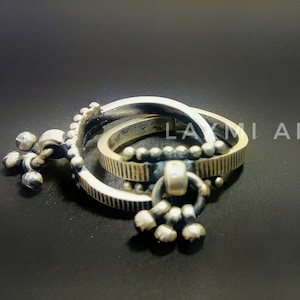 Puede incluir: Dos anillos de plata con un diseño decorativo. Los anillos están conectados por un pequeño amuleto de plata con tres cuentas redondas.