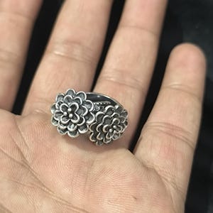 Puede incluir: Un anillo de plata con dos diseños de flores. Las flores son detalladas y tienen una superficie texturizada.