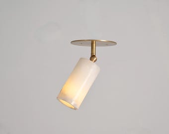 May include: A modern ceiling spotlight fixture. It features a cylindrical, off-white shade and a brass-colored mounting plate and arm. The light is angled downward, illuminating the interior of the shade. The fixture is designed for indoor use.