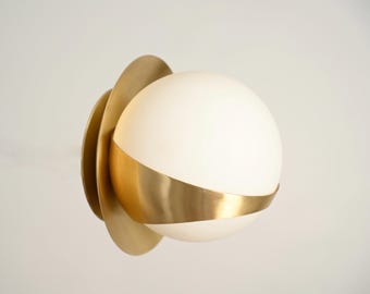 May include: A modern wall sconce featuring a spherical, frosted glass globe encircled by a wide, brass band. The globe is set against a backdrop of overlapping, petal-shaped brass elements, creating a layered, artistic design. The fixture is designed for interior lighting.