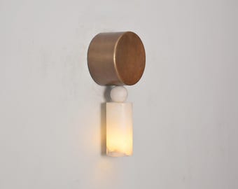 May include: A modern wall sconce with a cylindrical bronze-colored top and a white, rectangular, illuminated bottom. A small white sphere connects the two sections. The sconce is mounted on a white wall.