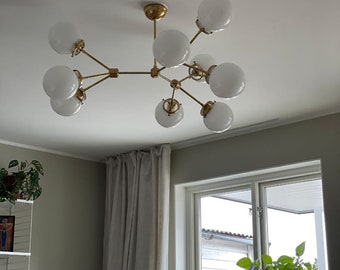 May include: A modern chandelier with a brass finish and multiple white globe-shaped glass shades. The fixture is mounted on a white ceiling and is designed with a branching, sputnik-style aesthetic. The room has a neutral color palette.