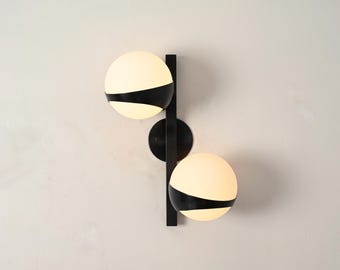 May include: Modern wall sconce featuring two spherical, off-white glass shades with a black band, mounted on a black metal frame. The fixture is designed with a vertical bar and a circular base, offering a contemporary lighting solution.