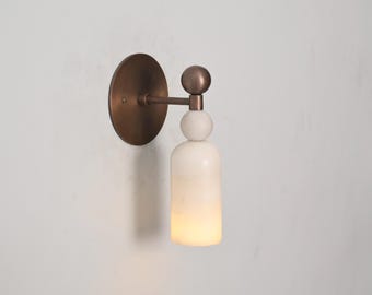 May include: A wall-mounted sconce with a circular bronze backplate and a cylindrical, off-white shade. The shade is topped with a small, round, bronze sphere and a white sphere. The sconce is designed for indoor lighting.