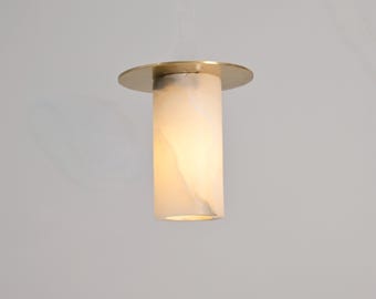 May include: A cylindrical, off-white lamp with a brass-colored circular top. The lamp's body is translucent, allowing light to pass through. The design is minimalist and modern, suitable for various interior styles.