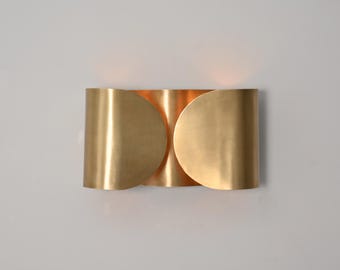 May include: A modern, gold-colored wall sconce with a unique design. The sconce has a curved, semi-circular shape with a warm, inviting glow emanating from within. The fixture is mounted on a neutral-colored wall.