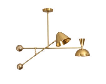 May include: A modern gold-toned ceiling light fixture with three light heads. The fixture has a central rod with a circular base, and three arms extending outwards, each with a different shaped light head. The light heads are cone, dome, and sphere shaped.