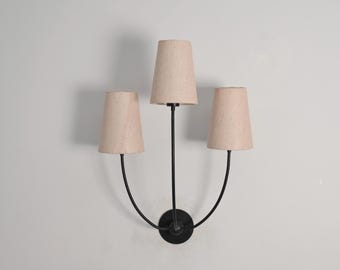 May include: A three-light wall sconce with a black metal frame and beige fabric shades. The sconce has a circular base and curved arms supporting the shades. The shades are cone-shaped and evenly spaced.