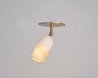 May include: A modern ceiling spotlight with a white, cylindrical shade and a brass-colored mounting plate and arm. The light is angled downward, illuminating the interior of the shade. The fixture is designed for indoor use.
