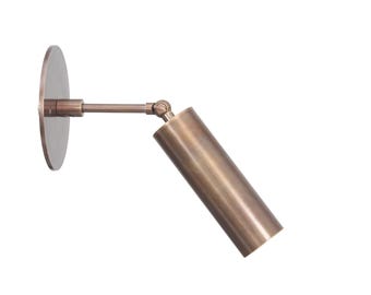 May include: A wall-mounted spotlight fixture with a circular backplate and a cylindrical head, both finished in a brushed bronze tone. The lamp features a horizontal arm connected to the light head by a swivel joint for adjustable positioning.