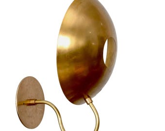 May include: A brass wall sconce with a curved arm and a large, oval-shaped shade. The shade has a circular opening and a polished finish. The sconce is mounted on a round base.