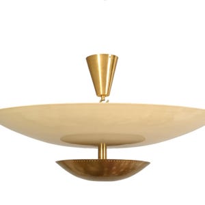 Op de afbeelding: Een plafondlamp met een grote, ondiepe, gebroken witte schotelvormige kap die hangt aan een conische messing plafondkap. Een kleinere, geperforeerde messing schaal is onder de hoofdkap bevestigd door een centrale messing stang.